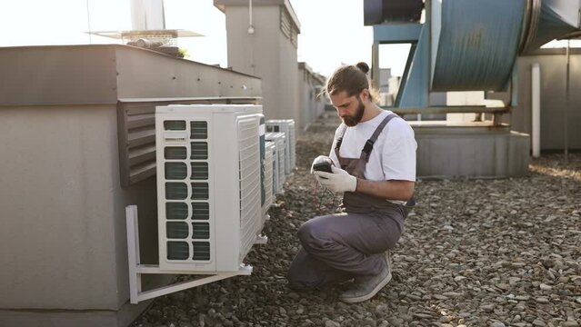 Concentrated man squatting down and holding voltage tester while applying to air conditioner. Builder man closed in overalls performing work and checking serviceability of cooling system.