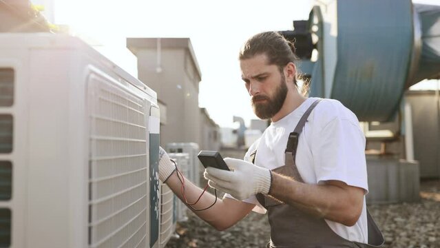 Side view of professional builder sitting in front of air conditioner and using measuring device on roof of factory. Caucasian focused male with braided hair testing voltage of climate control unit.