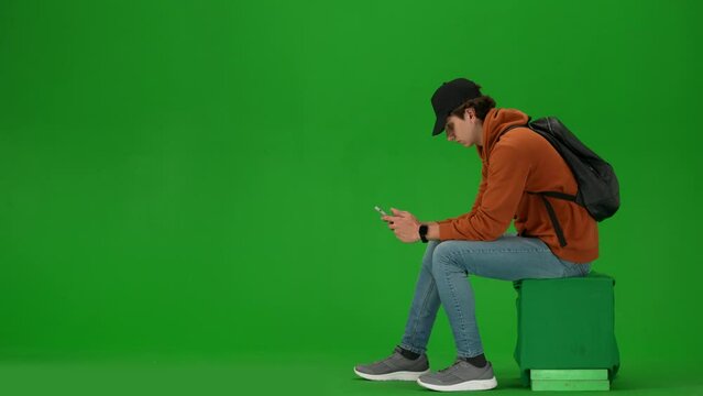 Portrait of traveler isolated on chroma key green screen background. Man in cap sitting texting on smartphone, taking selfie on camera app.