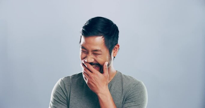 Funny, face and man laughing in a studio for comic or silly joke in conversation with happiness. Smile, portrait and young Asian male model with goofy facial expression isolated by gray background.