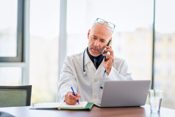 A mid aged male doctor sitting in doctor's office and talking to his patinet on the phone