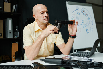 Bald mature male coach of military training sitting by workplace and holding gun while showing it...