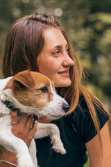 Close-up of a young positive woman hugging friendly Jack Russell Terrier dog, enjoying happy moments together while walking in the park on a summer day.