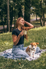A young positive woman walks with her dog in the park.A woman drinks tea from a thermos.A Jack Russell Terrier dog is sitting next to her.