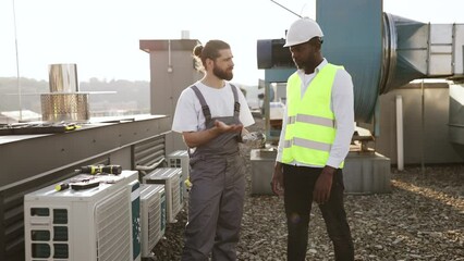 Caucasian bearded man in overalls standing and pointing to air conditioner while afro supervisor evaluating work. Two repairmen assessing quality of installed devices on roof of factory.