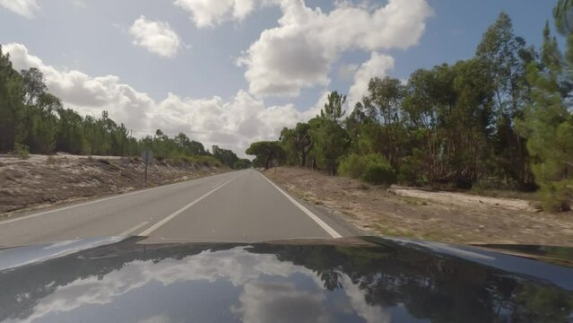 First person view, FPV, from dashcam of car driving along the Alentejo Coast in Portugal, passing cork oak trees and sand dunes. Road trip video in POV, with blue sky and clouds on an empty road