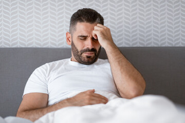 Unhappy man lying in bed touching head having migraine indoor