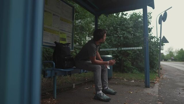 Tired and stressed man sits at bus stop and waits long time for transport, looking at watch and road. Passenger uses phone at station in Germany, Munich. Awaiting for bus for longer duration. 