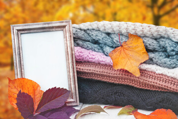 Hello autumn. A stack of knitted winter clothes, colorful autumn leaves and an old empty picture frame with copy space on a table over abstract blurred background.