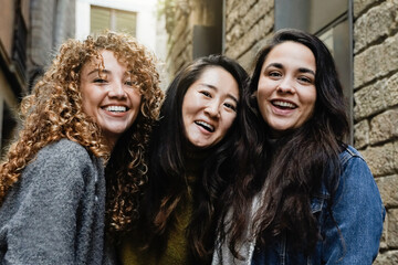 Three young women smiling at camera outdoors in city. Multiracial friends taking selfie picture...