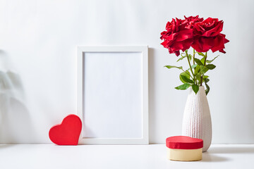 Mockup with a white frame and red roses in a vase, red heart  on a white table. Empty poster frame...