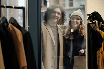 Happy young couple in stylish casualwear looking through boutique display at new seasonal fashion...