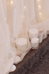 Obraz na płótnie Canvas Unlit white candles in glass containers, set on a gray carpet. A sheer white curtain with small lights forms the backdrop, creating a soft, romantic ambiance. Concept: Cozy home decor. 