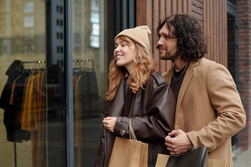 Happy young woman and her boyfriend with paperbags looking through shop window at new seasonal...