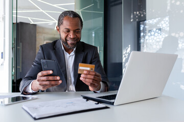 Smiling african american man working in office at desk, holding credit card and using mobile phone.