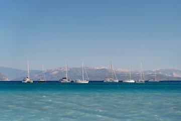 A tranquil line-up of sailboats floating on the clear blue sea with distant mountains under a clear...
