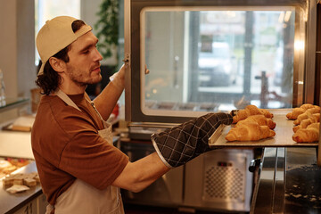 Professional male baker looking at tray with croissants taking it out from oven at kitchen in cafe