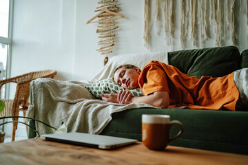 Young woman sleeping on comfortable sofa with cushion at home near laptop and mug