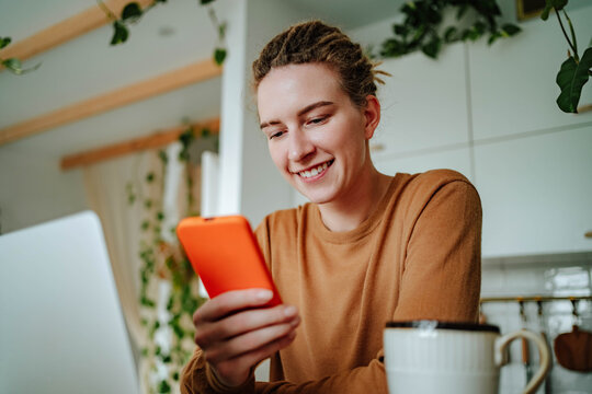 Smiling young woman using smartphone at kitchen table with laptop in daylight