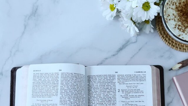 Hand turning pages on open holy bible book with cup of coffee and flowers on white marble background. Top view. Christian education, studying Scriptures at home concept.