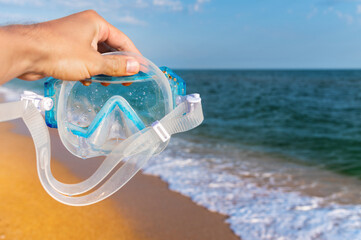 outstretched male hand holds a snorkeling mask on the sea of sea waves and a golden sandy beach....