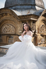 Fototapeta premium bride in a white dress sits on a ledge with an old building in the background. Young Caucasian woman straightens her hair at a wedding in an ancient city