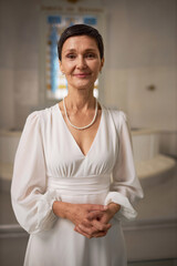 Vertical portrait of smiling senior woman as happy bride standing at altar in church and looking at...