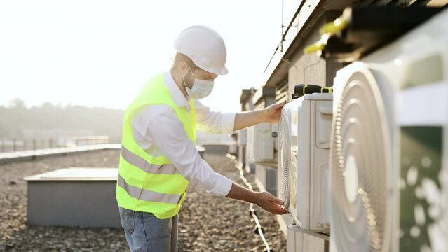 Professional worker of plant wearing dust mask crouching opposite air conditioner and providing inspection on fresh air. Caucasian bearded man checking attachment of device to wall in urban roof.