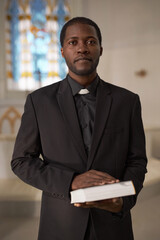 Vertical waist up portrait of Black young man as priest holding Bible standing at altar in church