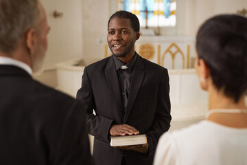 Waist up portrait of smiling Black young man as priest officiating wedding in church and holding...