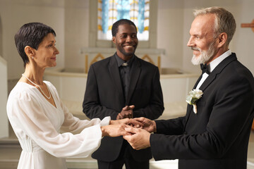 Side view portrait of loving senior couple getting married at church altar with priest officiating...