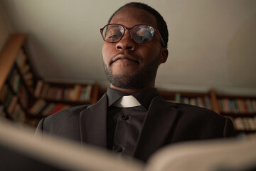 Low angle portrait of Black man as priest reading Bible in church office preparing for Sunday...