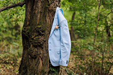 The groom's blue jacket with a boutonniere hangs on a tree in the forest. Wedding photography.