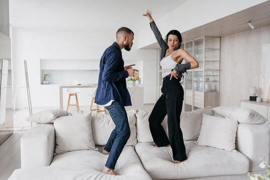 Young couple dancing on couch at home celebrating relocation, new apartment. Happy newlyweds enjoying vacations at hotel. Playful man having fun with girlfriend.