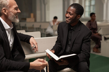Portrait of young Black man as smiling priest talking to senior man after Sunday service in catholic church, copy space