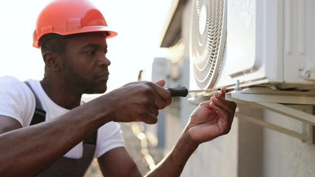Focused african craftsman wearing orange hard hat holding screwdriver while screwing bolt into air conditioner. Serious specialist repairing broken device and performing work on rooftop.