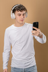 One man caucasian young male stand studio shot on beige background use mobile phone smartphone with headphones listen music send messages texting or browse internet online app copy space