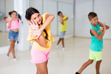 Portrait of cheerful preteen hispanic girl learning energetic dance moves with group of children in choreography class .