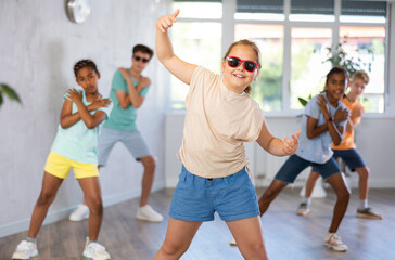 Fototapeta premium Dynamic little girl training Break-dance poses in dancehall with other attendees of dancing courses