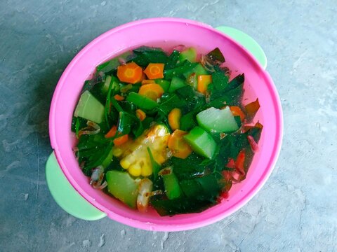 sayur bening or clear vegetables in a pink bowl on a gray background. Healthy vegetable soup filled with corn, carrots, katuk leaves or leaf vegetable, chayote and spring onions. nutritious and health