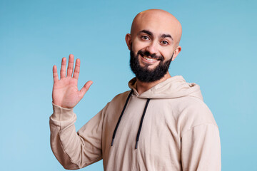 Cheerful arab man waving hi with hand and looking at camera. Happy young bald bearded person with...