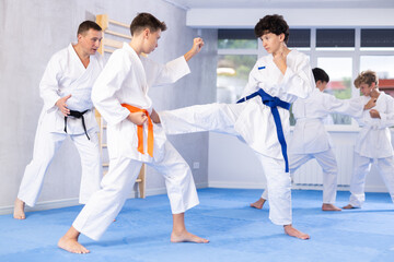 Concentrated teen boys in kimonos honing kicking techniques during kumite at karate training session under guidance of sensei