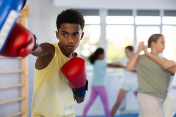 Foto op Canvas Motivated focused african american boy wearing boxing gloves working out with coach during group self-defense course, practicing punches on mitts.. © JackF