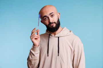 Thoughtful arab man holding pen and tilting head while looking at camera with pensive facial expression. Young bald bearded person planning, thinking and dreaming studio portrait