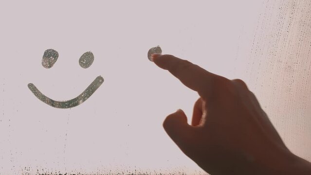 Woman's finger painted doodle two sad face and happy smile on window flooded with raindrops, concept of opposites
