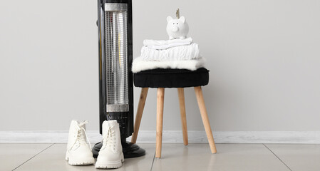 Electric radiator with winter boots, knitted sweaters and piggy bank near light wall in room