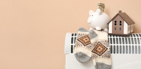 Electric radiator with knitted socks, figure of house and piggy bank near beige wall in room....