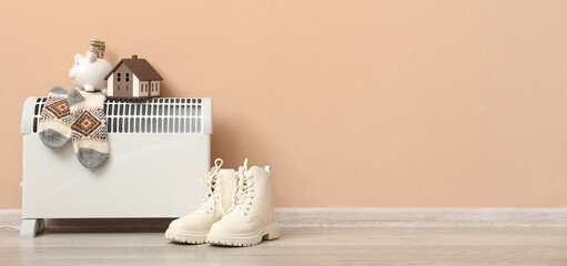 Electric radiator with warm socks, figure of house, boots and piggy bank near beige wall in room....
