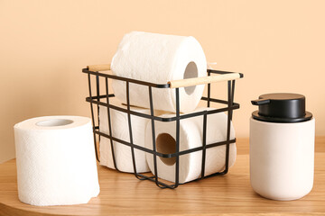 Basket with toilet paper rolls and bottle of soap on wooden table near color wall, closeup