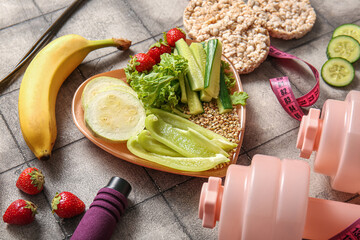 Plate with fresh healthy products and dumbbells on grey tile background, closeup. Diet concept